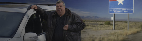 A man in a brown leather jacket stands beside a white vehicle in an open landscape. The jacket has a plush collar and a vintage design. A sign in the background reads,