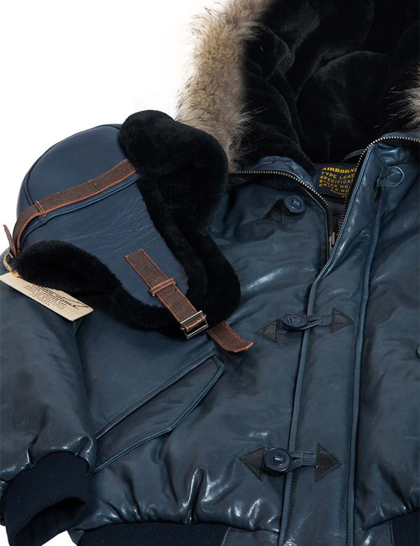 Men's navy blue trapper hat with brown straps, leather upper and sheepskin lining, placed on a short Alaska leather jacket with a hood