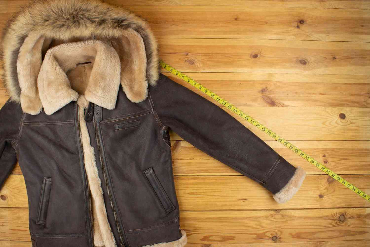 The image shows a brown shearling jacket with a fur-lined hood, placed on a wooden surface. A yellow measuring tape is stretched out next to the jacket, indicating its dimensions. The jacket features a zip closure and side pockets, with the fur lining visible along the collar, hood, and cuffs, highlighting the garment's warm and cozy design.