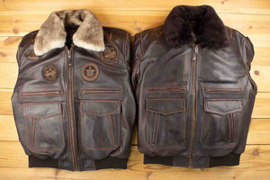 The image displays two dark brown leather jackets side by side on a wooden surface. Both jackets feature zip closures, multiple front pockets, and fur-lined collars—one in a lighter beige fur and the other in darker brown fur. The jacket on the left has various embroidered patches, giving it a vintage, military-inspired look, while the one on the right is more minimalistic without any patches. The rugged texture and detailed stitching highlight the craftsmanship of these jackets.