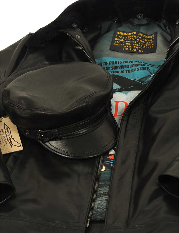 A men's black leather cap with a strap is placed over a black leather jacket with a fur collar.