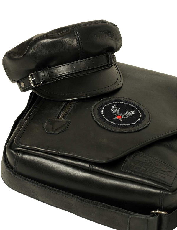 A men's black leather cap with a strap is placed on top of a black leather bag with a patch.